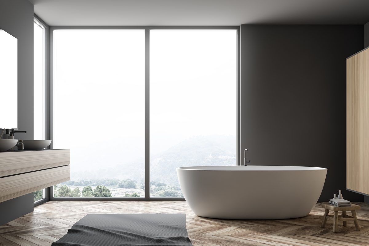 Bodentiefe Fensterfront in einem hellen Badezimmer mit freistehender Wanne und Blick auf bewaldete Hügel