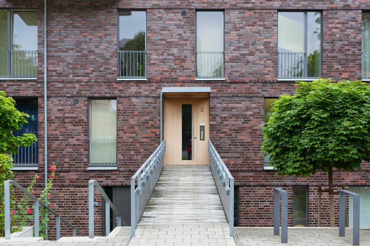 Gepflasterter Zuweg und ein Holzsteg mit Geländer führen auf die Eingangstür eines Mehrfamilienhauses mit Klinkerfassade