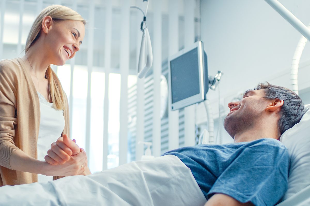 Frau am Krankenbett hält lächelnd die Hand eines Patienten im Krankenzimmer
