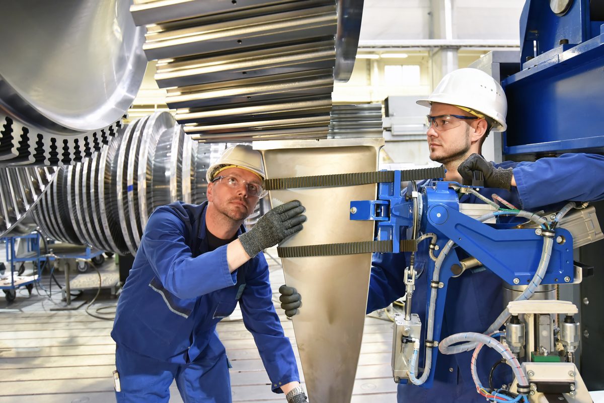 Zwei Monteure mit Schutzhelm und Schutzbrille setzen in einer Werkhalle ein Bauteil an einer Turbine an, links der offene Läufer mit seinen Schaufelkränzen