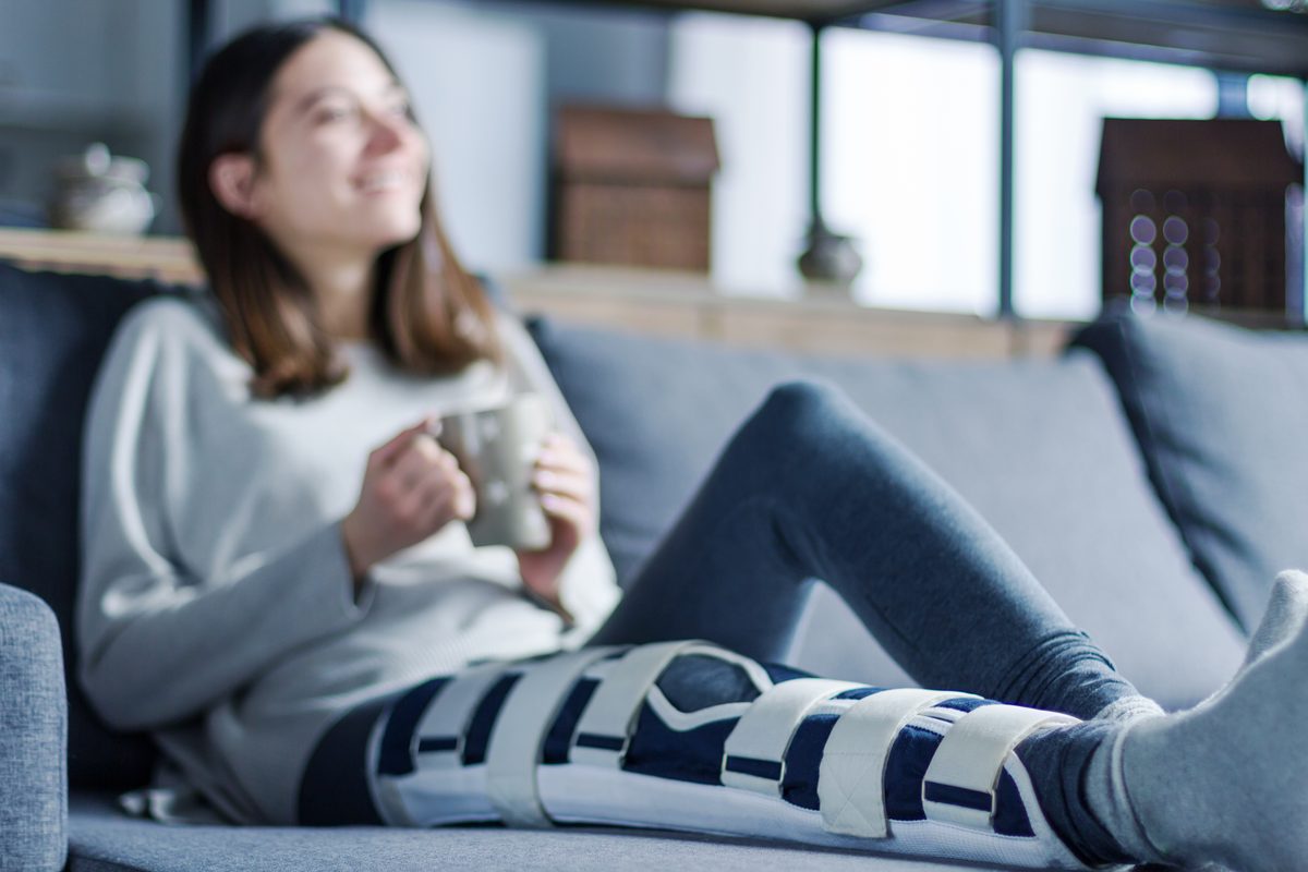 Junge Frau mit Knieschiene sitzt mit einer Tasse Tee auf dem Sofa