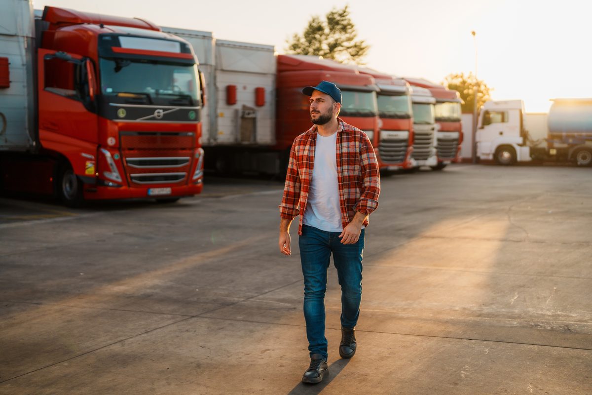Mann mit Schirmmütze geht über einen Betriebshof, dahinter eine Reihe geparkter Lkw im Abendlicht